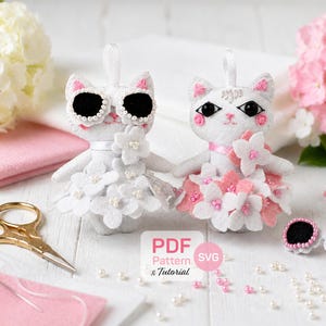 Puede incluir: Dos muñecos de gato de fieltro blanco con lazos para colgar, uno con gafas de sol negras y vestido de flores blancas, el otro con detalles florales rosas y blancos. Incluye tijeras doradas, cuentas sueltas y una etiqueta PDF Pattern & Tutorial SVG.