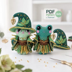 May include: Two felt plush figures, a cat and a frog, wearing dark green pointed hats with gold leaf charms and beaded skirts. The cat has light green fur and the frog is dark green. A PDF Pattern & Tutorial label is visible in the corner.