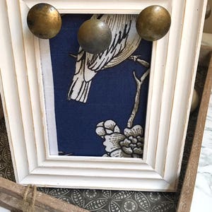 May include: A white picture frame with a blue fabric insert featuring a white bird and floral design. Three gold-toned metal knobs are attached to the frame.