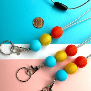 Op de afbeelding: Kleurrijke kralen lanyards met een karabijnhaak en sleutelring. De lanyards hebben een reeks blauwe, gele en rode bolvormige kralen. Een munt is inbegrepen ter referentie. De achtergrond is verdeeld tussen blauw en roze.