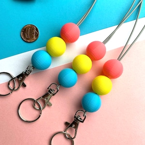 Op de afbeelding: Drie kralen lanyards met pastelkleurige houten kralen in roze, geel en blauw. Elke lanyard heeft een zilverkleurige sluiting en een grijs koord. Een munt ligt naast de lanyards ter vergelijking.