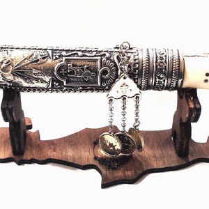 Puede incluir: Un cuchillo decorativo de plata y oro con mango de marfil, expuesto en un soporte de madera con forma de la isla de Chipre.