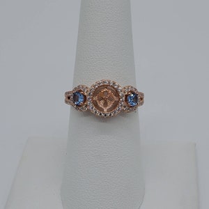 May include: A rose gold ring with a center stone setting surrounded by a halo of smaller stones. Two smaller blue stones are set on either side of the center setting.