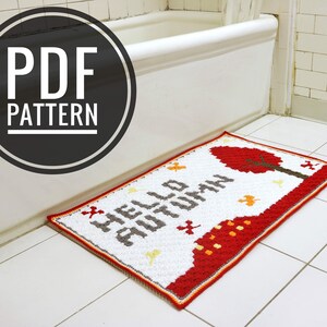 May include: A white crochet bath mat with the text "Hello Autumn" in gray and a red tree design. The mat is in a bathroom with white tile floors and a white bathtub.