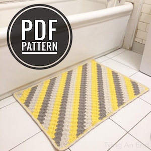 Puede incluir: Una alfombra de baño de crochet con un patrón de rayas diagonales en amarillo y gris. La alfombra está en un baño con una bañera blanca y un suelo de baldosas blancas. El texto "PDF PATTERN" está en un círculo negro en la esquina superior izquierda de la imagen.