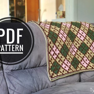Op de afbeelding: Een PDF-patroon voor een gehaakt deken met een ruitpatroon in tinten groen, bruin en roze. Het deken ligt op een grijze bank. De tekst "PDF PATTERN" staat in een cirkel aan de linkerkant van de afbeelding.