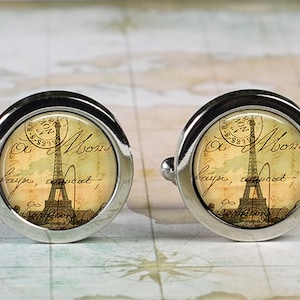 May include: Pair of silver-toned cufflinks with a vintage design. Each cufflink features a circular face with an image of the Eiffel Tower, script, and a postmark. The background is a sepia tone.