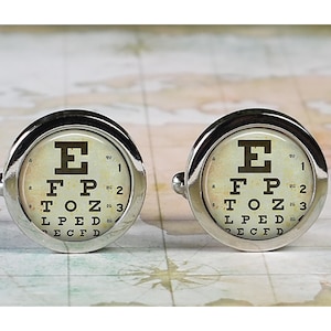May include: A pair of silver-toned cufflinks featuring a vintage-style eye chart design. The eye charts display the letters E, F, P, T, O, Z, L, P, E, D, and E, C, F, D. The cufflinks have a round face and a shiny finish.