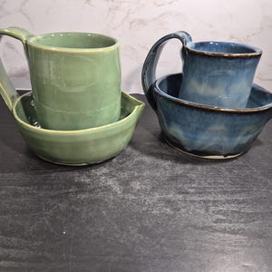 May include: Two sets of handmade ceramic pottery, each consisting of a mug and a matching bowl with a small pouring spout. One set is finished in a smooth sage green glaze, while the other set features a variegated blue glaze with dark brown edges.