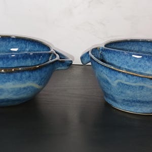 May include: Two blue ceramic bowls with handles. The bowls have a unique design with a divided interior and a textured, glazed finish. The rims are accented with a darker color. The bowls are placed on a dark surface against a white background.