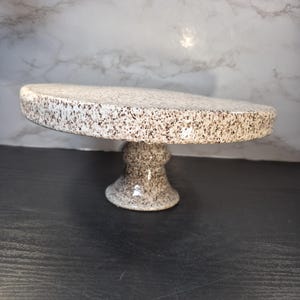 May include: A speckled ceramic cake stand with a round top and a pedestal base. The top and base are off-white with brown speckles. The stand is designed to display cakes, cupcakes, or other baked goods.