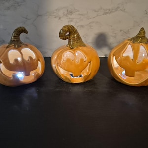 May include: Three orange ceramic jack-o'-lanterns with carved faces and brown stems. Each pumpkin features a hollow interior with a small, bright white LED light glowing from within, creating a warm illumination through the cutouts.