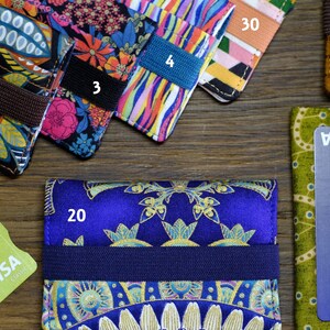 May include: A collection of ten colorful fabric card holders with different patterns. The card holders are arranged in a row on a wooden surface. The card holders are numbered 1 through 5 and 15, 20, 30. A green VISA credit card is also visible in the image.