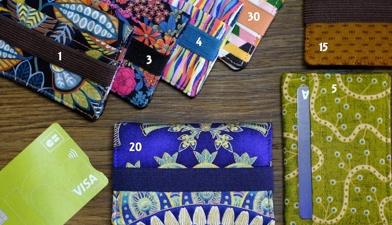 May include: A collection of ten colorful fabric card holders with different patterns. The card holders are arranged in a row on a wooden surface. The card holders are numbered 1 through 5 and 15, 20, 30. A green VISA credit card is also visible in the image.