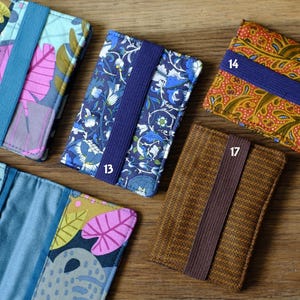 May include: Six different fabric card holders with elastic bands. The card holders are in various patterns and colors, including blue, brown, orange, and pink.