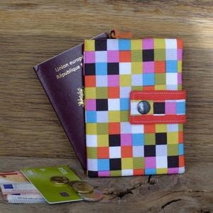 Peut inclure: Un porte-passeport coloré avec un motif patchwork de carrés multicolores. Une sangle rouge avec un bouton noir sécurise le porte-passeport. Un passeport et de la monnaie sont visibles. Le passeport indique 'Union européenne'.
