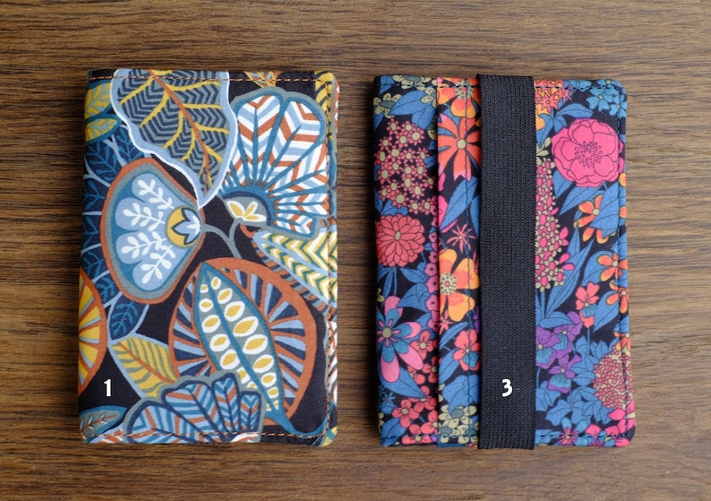 May include: Two fabric card holders with floral patterns. The holder on the left has a blue, green, and brown floral pattern. The holder on the right has a pink, orange, and blue floral pattern with a black elastic band.