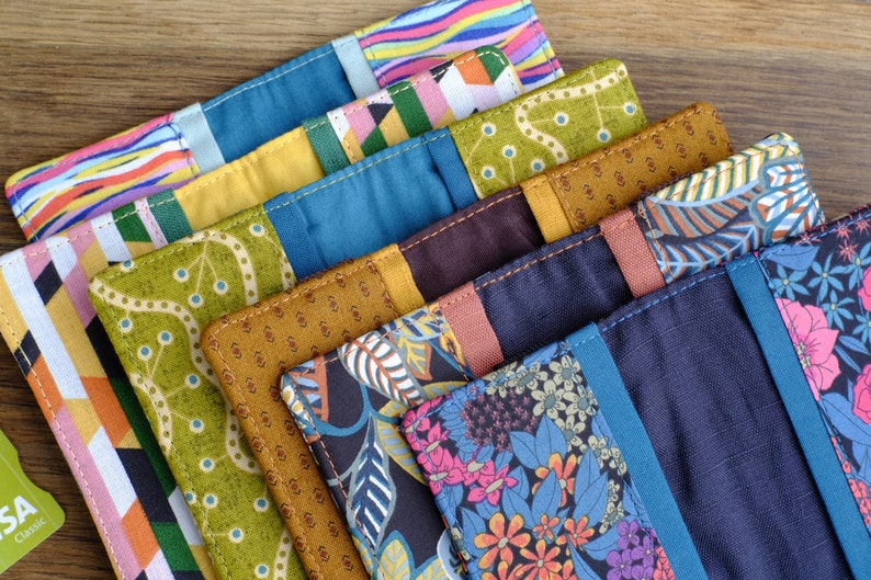 May include: A stack of eight colorful fabric pouches with different patterns, including stripes, geometric shapes, and floral designs. The pouches are made of a variety of fabrics, including cotton and linen. They are all sewn with a contrasting thread.