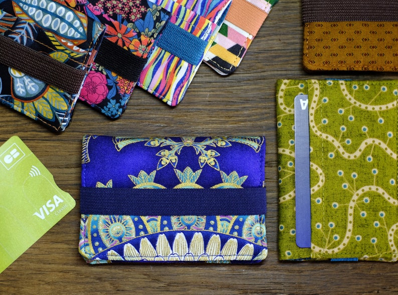May include: A blue and gold patterned card holder with a navy blue elastic band. The card holder is made of fabric and has a paisley design.