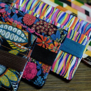 May include: Four colorful fabric wallets with different patterns. Each wallet has a brown, black, or blue strap.