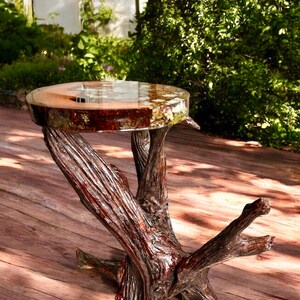 May include: A unique side table with a round, clear resin top, showcasing embedded natural elements. The table's base is crafted from intricately textured, dark brown wood, resembling a tree trunk. The table stands on a wooden deck, surrounded by greenery.