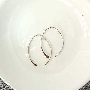 May include: A pair of silver-colored, minimalist hoop earrings. The earrings are curved, with a teardrop shape at the bottom. They are displayed on a white, round plate.