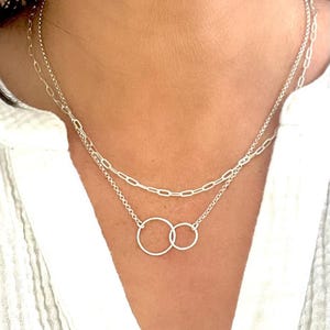 May include: A silver necklace with two layers. The top layer has a chain link design, and the bottom layer features two interlocking circles. The necklace is worn against a white shirt.