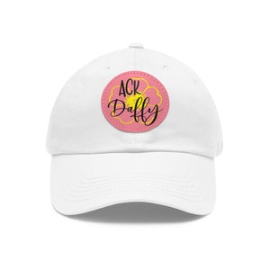 Può includere: Cappellino da baseball bianco con una toppa a forma di fiore rosa che dice "ACK Daffy" in lettere nere.