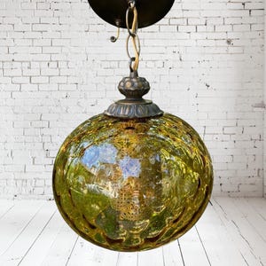 Pode incluir: Uma luminária pendente vintage em forma de globo. A sombra de vidro é de cor verde-oliva texturizada, com um padrão decorativo. A luminária tem uma copa e corrente de metal cor de bronze.