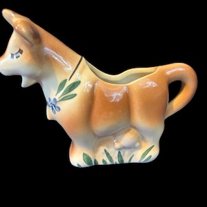 May include: A brown ceramic creamer shaped like a cow with blue and green floral details. The creamer has a handle and a spout.