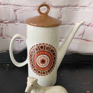 May include: A white ceramic coffee pot with a brown lid and a geometric design in shades of orange, brown, and white. A white ceramic dog figurine is lying in front of the coffee pot.