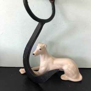 May include: A black metal hook with a wooden handle. The hook is curved and has a sharp point. A white porcelain dog figurine is lying in front of the hook.