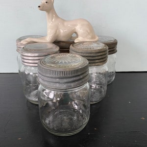 Op de afbeelding: Zes vintage glazen potten met zilveren deksels. De potten zijn in een cirkel gerangschikt met een wit porseleinen beeldje van een windhond erop.