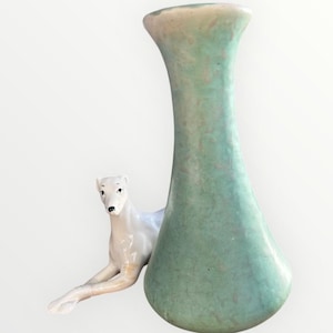 Puede incluir: Un jarrón alto de color verde claro con la parte superior ensanchada y una base redondeada. Una figura de perro de cerámica blanca está colocada junto al jarrón. El jarrón tiene una superficie texturizada y un aspecto vintage.
