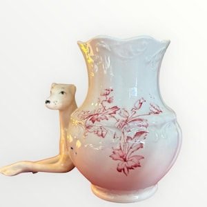 May include: A white and pink ceramic vase with a red floral pattern. A ceramic dog figurine is positioned to the left of the vase. The vase has a scalloped rim and a rounded base.