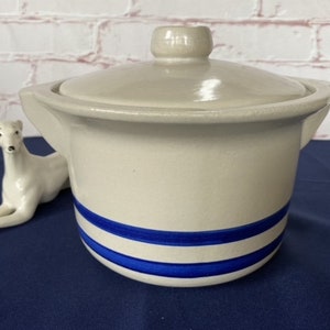 May include: A cream-coloured ceramic casserole dish with a matching lid and two side handles. The exterior features two horizontal blue stripes encircling the lower portion of the pot. The surface has a smooth, glossy finish throughout the piece.
