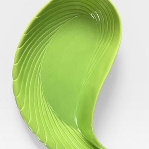 May include: A lime green ceramic dish with an asymmetrical, teardrop-like shape. The surface features a series of raised, curved ridges that follow the contour of the left side, leading to a smooth, concave centre and a pointed bottom tip.