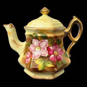 May include: A small, yellow teapot with a gold lid and handle. The teapot is decorated with pink and white flowers and green leaves.