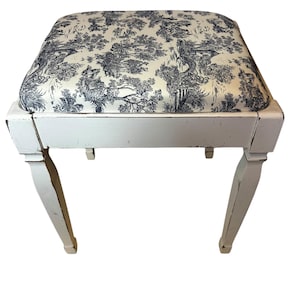 Puede incluir: Taburete de madera blanco con un cojín estampado en toile azul y blanco. El taburete tiene cuatro patas torneadas.