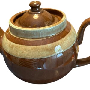 May include: A brown ceramic teapot with a rounded body, spout, and handle. The teapot features horizontal bands of brown and beige. The lid has a small knob. A classic kitchenware item.