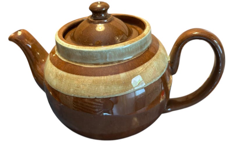 May include: A brown ceramic teapot with a rounded body, spout, and handle. The teapot features horizontal bands of brown and beige. The lid has a small knob. A classic kitchenware item.