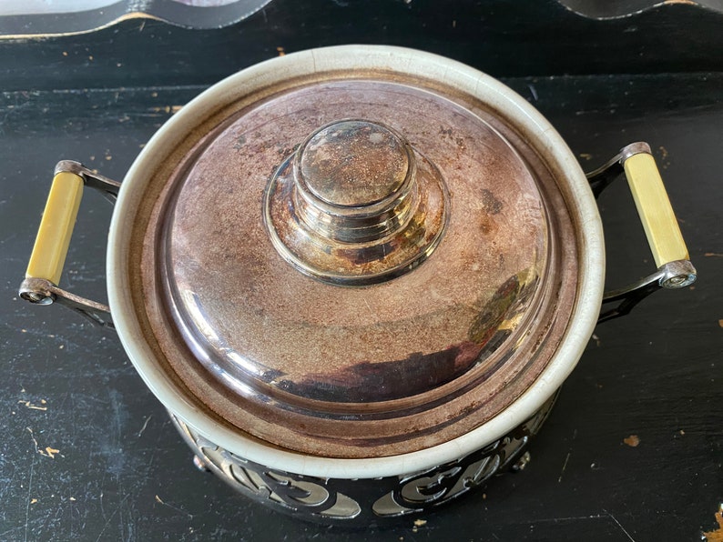 K&ouml;nnte beinhalten: Eine silberne Chafing Dish mit einer wei&szlig;en Porzellan-Innenauskleidung und einem dekorativen Metallsockel. Der Deckel hat einen silbernen Knopf und ist leicht ge&ouml;ffnet.