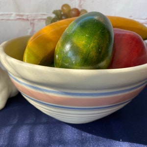 Puede incluir: Un tazón de cerámica blanco con rayas rosas y azules, lleno de una variedad de frutas, incluyendo una pera verde, un plátano amarillo y una manzana roja. El tazón está sentado sobre una superficie azul.