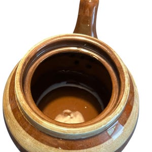 May include: A brown and tan ceramic jar with a handle. The jar has a round shape with a wide opening and a dark brown interior. The exterior features alternating bands of brown and tan.