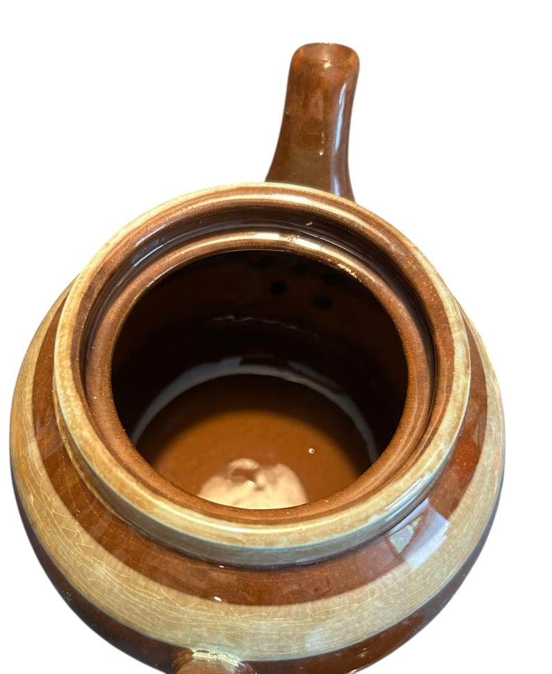 May include: A brown and tan ceramic jar with a handle. The jar has a round shape with a wide opening and a dark brown interior. The exterior features alternating bands of brown and tan.