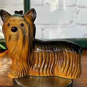 Może przedstawiać: Ceramiczna figurka psa rasy Yorkshire Terrier. Pies jest brązowy i czarny z błyszczącym wykończeniem. Figurka stoi na srebrnej podstawie.