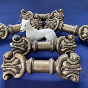 May include: Five ornate gold-toned metal drawer pulls with intricate designs. The pulls are arranged in a semi-circle with a white porcelain figurine of a dog in the center.