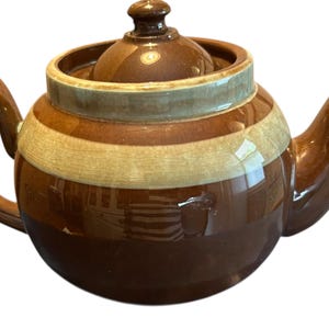 May include: A brown ceramic teapot with a lid and handle. The teapot has a rounded body with horizontal stripes in shades of brown and cream. The spout and handle curve gracefully.