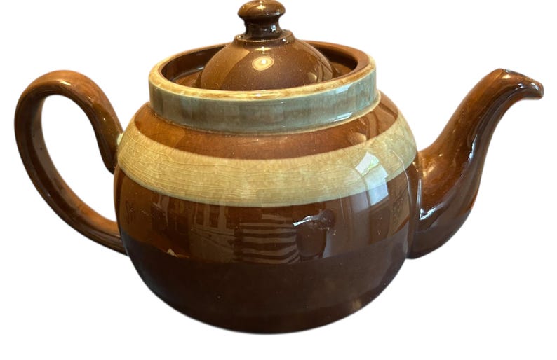 May include: A brown ceramic teapot with a lid and handle. The teapot has a rounded body with horizontal stripes in shades of brown and cream. The spout and handle curve gracefully.