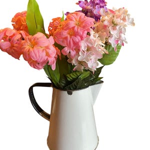 Peut inclure: Un pichet en émail blanc avec une poignée noire rempli d'un bouquet de fleurs artificielles roses, violettes et blanches.